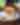 A juicy cheeseburger topped with crispy bacon sits on a white plate with pickles, set on a wooden table. A refreshing drink is blurred in the background.